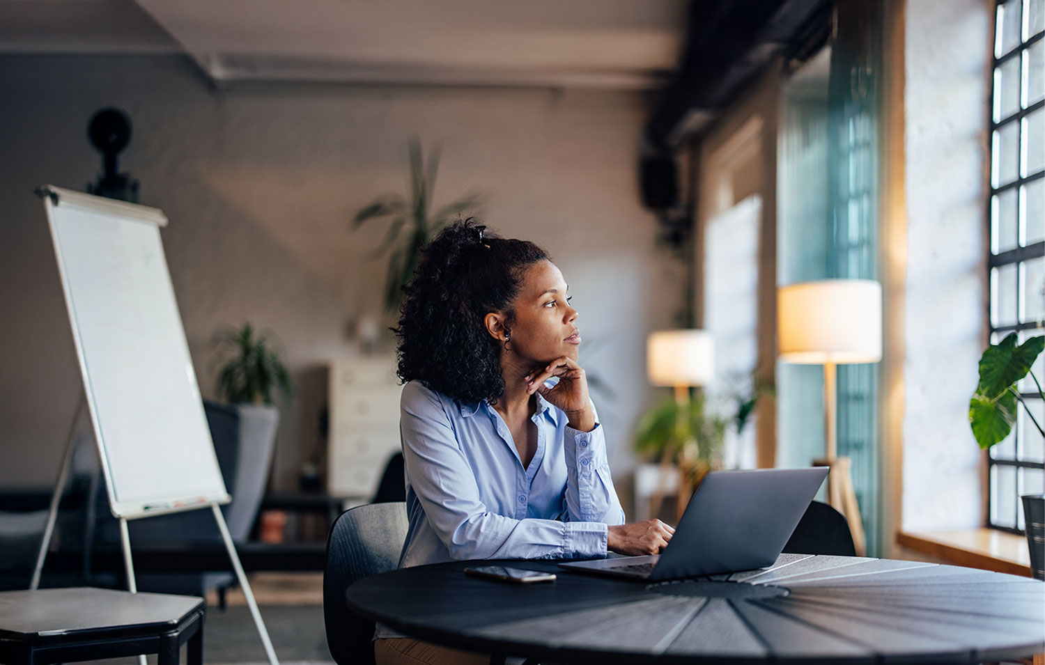 Woman gazing thoughtfully out the window while working on a laptop in a modern, bright office.
