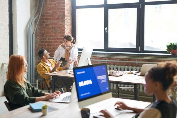 Modern open-plan office space with young professionals working at shared desks in a bright loft-style environment with exposed brick walls.