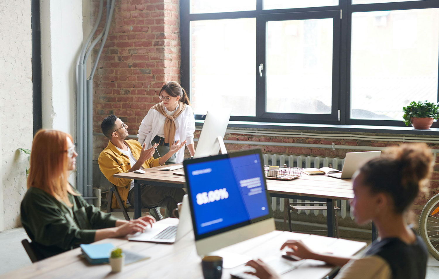 Modern open-plan office space with young professionals working at shared desks in a bright loft-style environment with exposed brick walls.